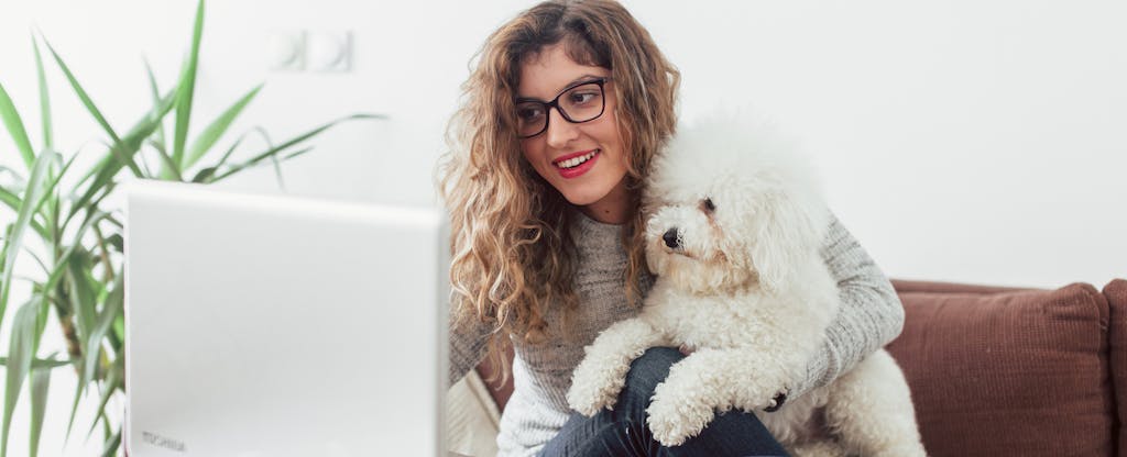 Smiling young woman holding a small white dog and typing on her laptop at home.