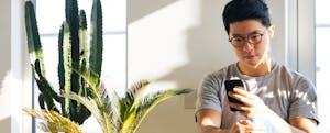 Young man with smartphone, sitting on the floor at home with houseplants