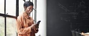 Woman looking at her phone and smiling while at the office, reading about get paid early apps