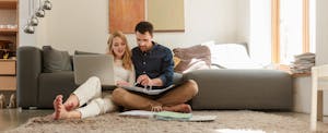 Couple seated on the floor beside a sofa research down payment assistance programs on a laptop.