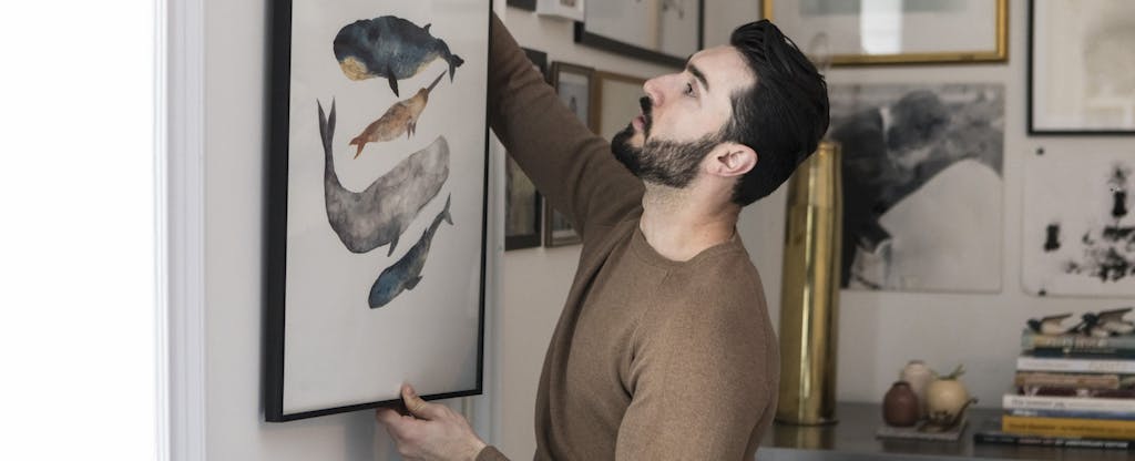Man staging home carefully hangs a painting of whales alongside other artwork.