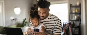 Father holds a toddler while checking his credit score on his phone in preparation to buy a house.