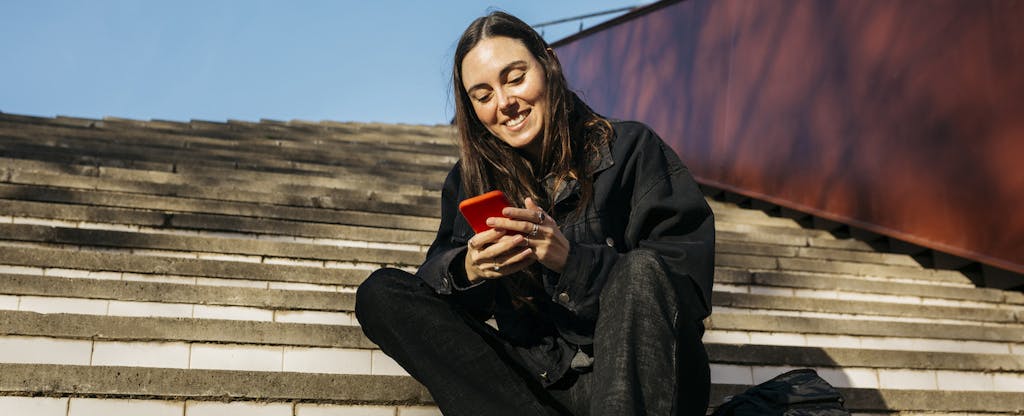 A smiling woman looks at her mobile device while sitting on a staircase in the sun.