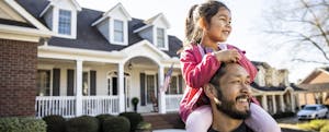 A young girl is sitting on her father's shoulders, in the front yard of their house.