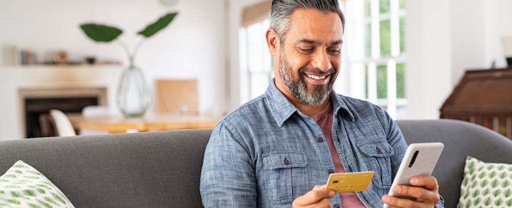 A smiling man sitting on a sofa looks at his credit card while holding his mobile phone..