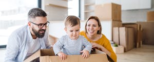 A couple in their new home smile as their toddler plays in an empty moving box.