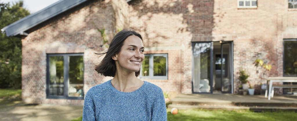 A smiling woman stands on the lawn in front of her brick home.