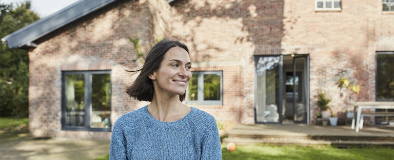 A smiling woman stands on the lawn in front of her brick home.