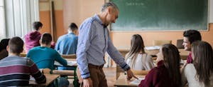 A teacher smiles as he stands next to a student's desk and assists them with a question.