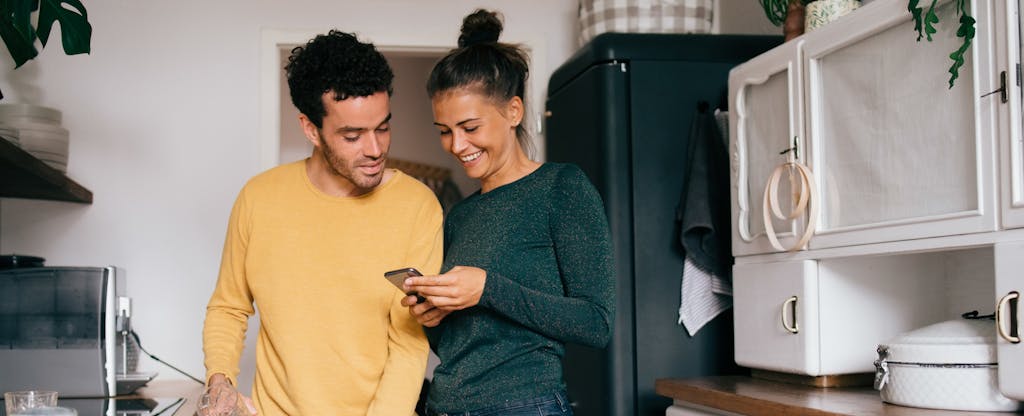 Two people standing together in their kitchen, smiling and looking at a cellphone together
