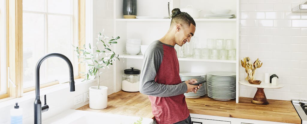 Young man standing alone in his kitchen, smiling as he reads on his phone about how to get overdraft fees refunded