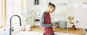 Young man standing alone in his kitchen, smiling as he reads on his phone about how to get overdraft fees refunded