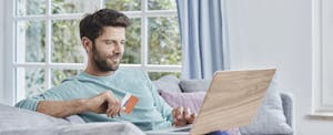 A smiling man seated on a sofa holds his laptop and a credit card while shopping online.