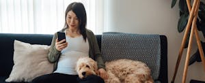 Young woman sitting on her sofa with her dog, looking at her automatic payments on her mobile phone
