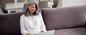 A woman seated on a sofa thoughtfully reads a review of the upgrade bitcoin card on her laptop.