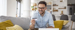 A smiling man wearing glasses holds his Upgrade Cash Rewards Elite Visa card while using a laptop.