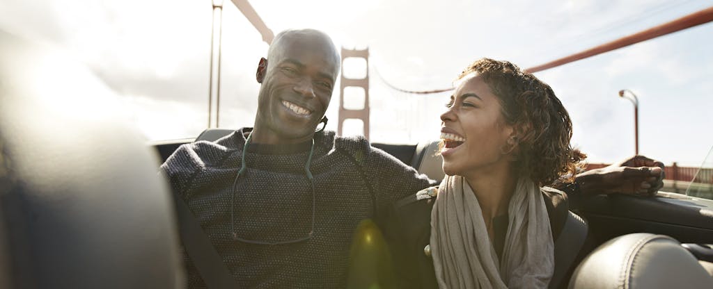 Man and woman sitting together in the front seat of their convertible, smiling as they drive across the Golden Gate Bridge