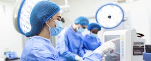 An anesthesiologist in blue scrubs and a surgical mask looks at a monitor.