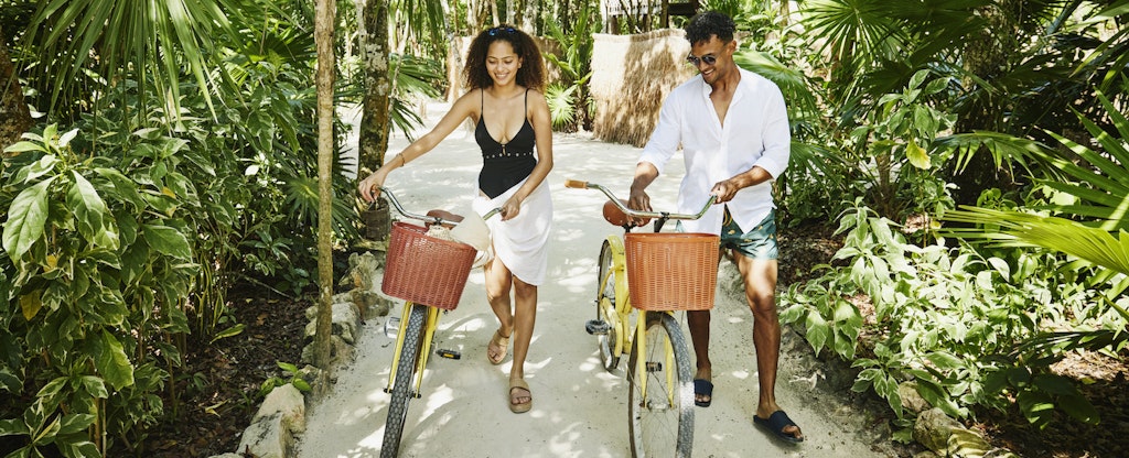 A smiling couple walk their bicycles along a sandy path surrounded by palms.