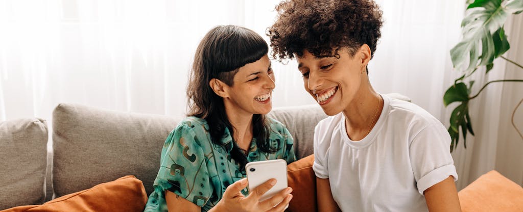 A couple seated on a sofa smile while using a cellphone to look at the Empower app together.