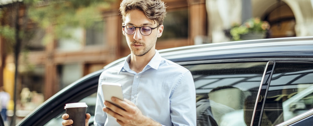 Man standing by his car holding coffee and looking at his phone