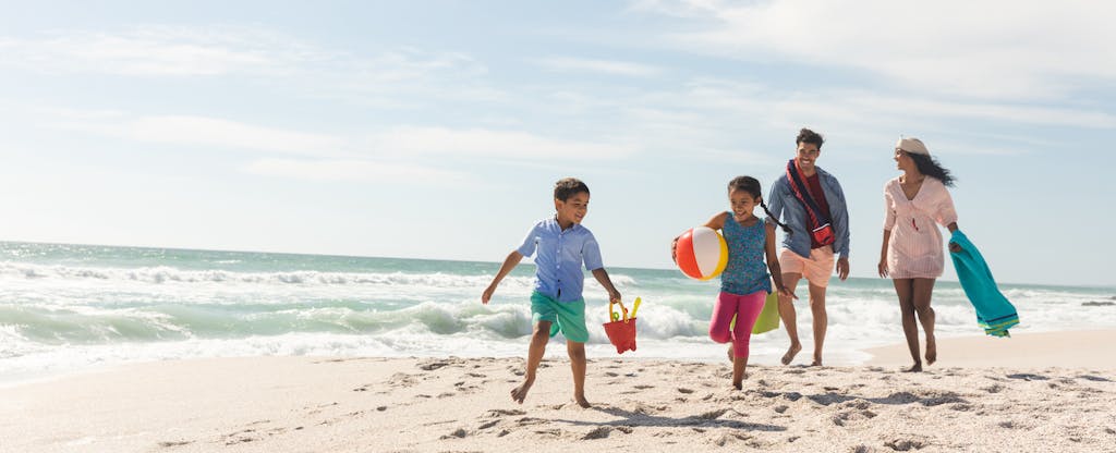 Parents and two young children carrying beach toys walk along the shore together.