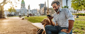 A man seated on a park bench next to his golden retriever smiles as he looks at his smartphone.