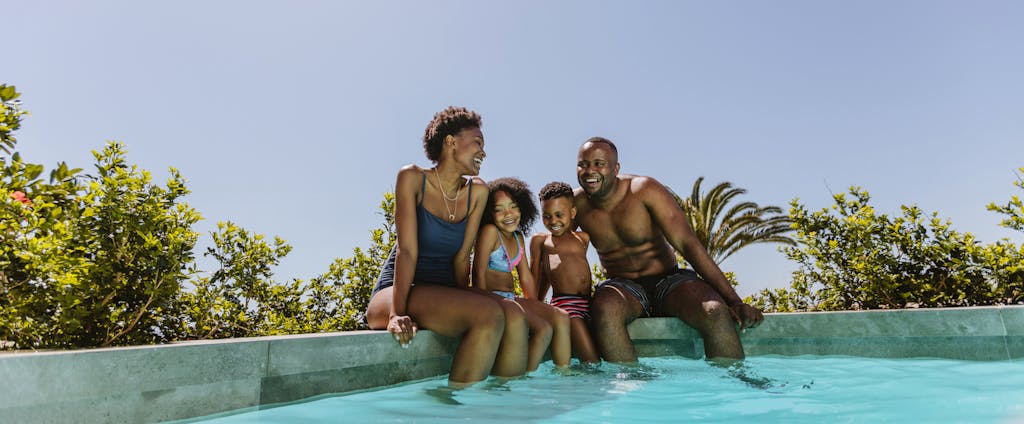 A smiling family sits on the edge of a pool dangling their feet in the water.