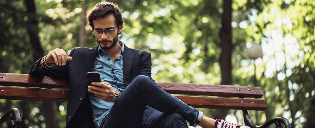 A man seated on a park bench looks thoughtfully at his smartphone.