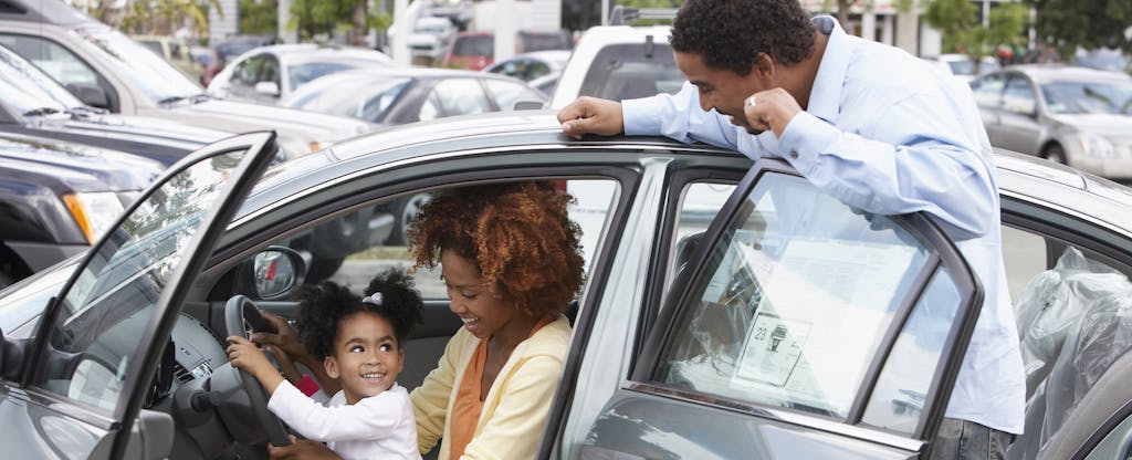 A father looks on as a mother and daughter sit in the driver's seat of a new car at an auto dealership.