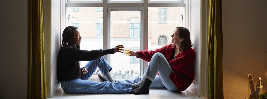 Two roommates sitting in the windowsill of their new apartment, smiling and toasting to their new place