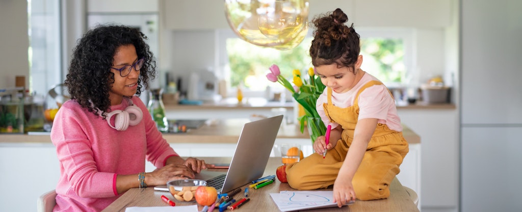 Woman sitting at her kitchen table, working on her laptop, while her child sits across from her, drawing