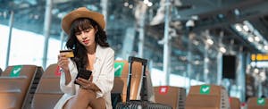 A woman wearing a straw hat and holding a credit card sits next to her luggage at an airport and uses her smartphone to review her American Airlines elite status benefits.