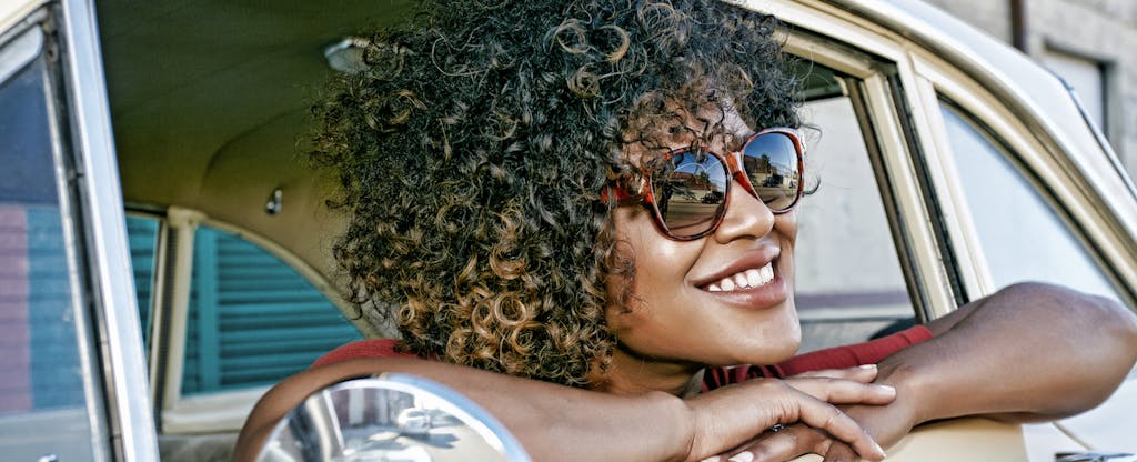 Young black woman smiling from the driver's side window of her classic car