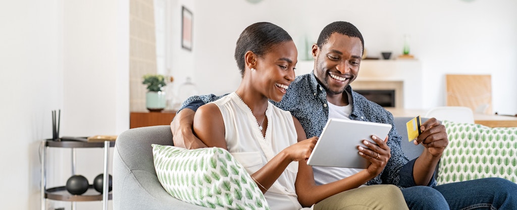 A smiling couple sit on their sofa and use a tablet to review redemption options through amex rewards.