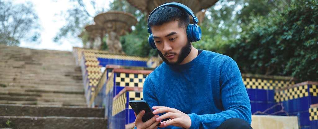 A man wearing a blue sweater and headphones sits on a park staircase and uses his smartphone to look into the upcoming Apple Pay Later service.