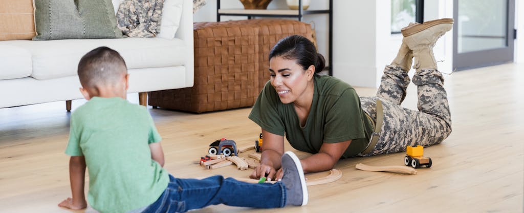 A mother wearing cargo military pants plays with her young son on the floor using wooden cars.