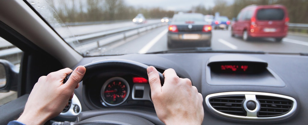 A close up of a pair of hands on a car steering wheel with a highway and other cars visible through the windshield.
