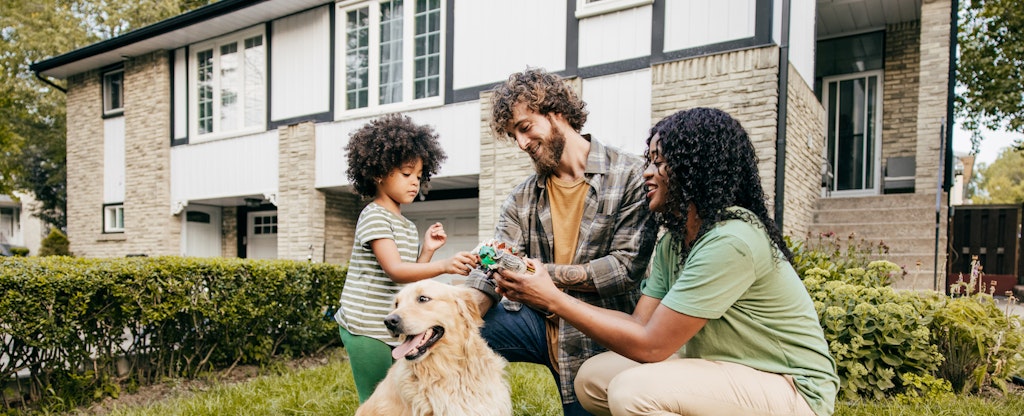 Parents and their young child play with dinosaur toys in the front yard of their home while their golden retriever sits nearby.
