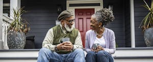 A couple smile at one another while having coffee on the porch steps of their home.