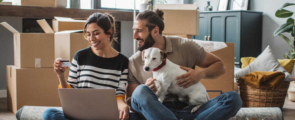 A couple sit on the floor of their home, one person holds their dog while the other holds a laptop and their Aven HELOC card.
