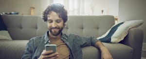 Man sitting in his living room, on the floor in front of his couch, looking at a Cleo cash advance on his mobile phone