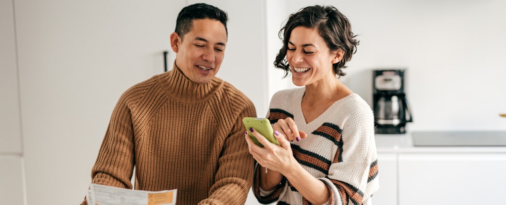 A couple review their finances together in the kitchen and smile while using a smartphone to read about the new credit scoring models for mortgages.