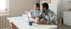 A man wearing military fatigues and his spouse sit at their dining room table and use a laptop to look into Pioneer military loans.