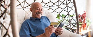 A seated man smiles while holding his smartphone and his sofi credit card.