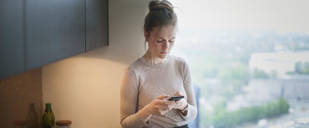 A woman stands near a window in her home while using a smartphone to read about auto insurance companies that do not use credit scores.