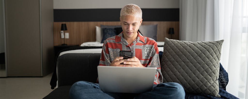 Young man sitting cross-legged on his bed using his smartphone and laptop.