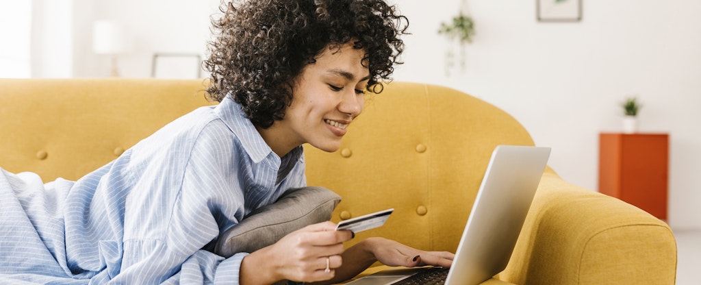 A smiling woman lying on a yellow sofa holds one of her premium rewards cards while using a laptop to review all its benefits.