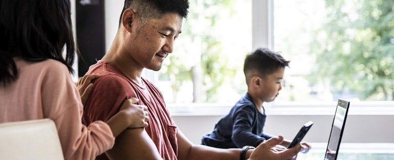 A man seated at a table in front of a laptop uses his smartphone while his two young children play alongside him.