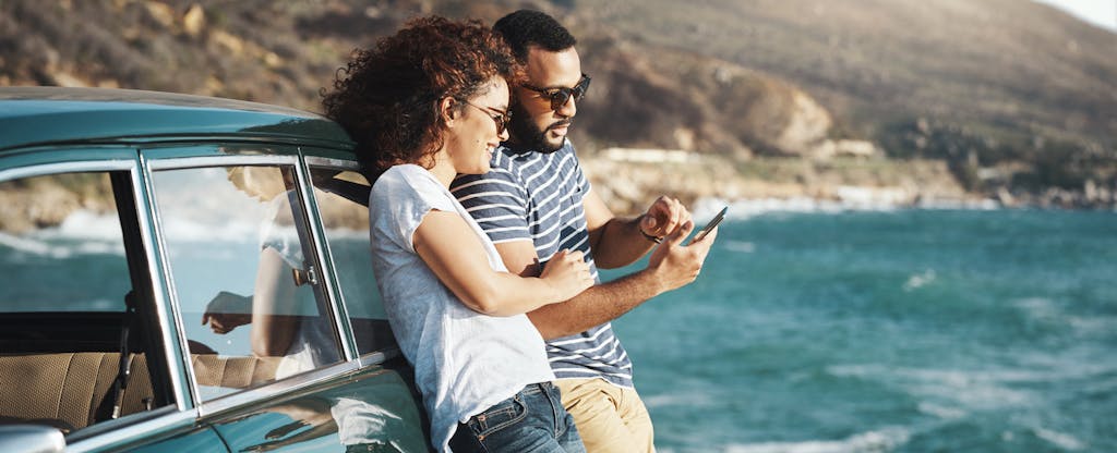 A couple lean against their parked car while using a smartphone to look into states that allow nonresident vehicle registration.